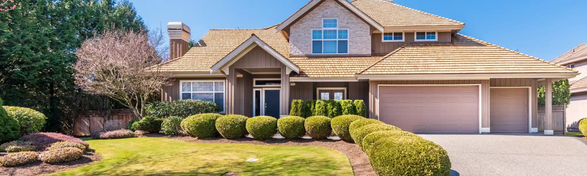 A large two-story suburban home with a cedar shake roof, board-and-batten siding, a double garage, and manicured landscaping including rounded shrubs and a green lawn on a sunny day.