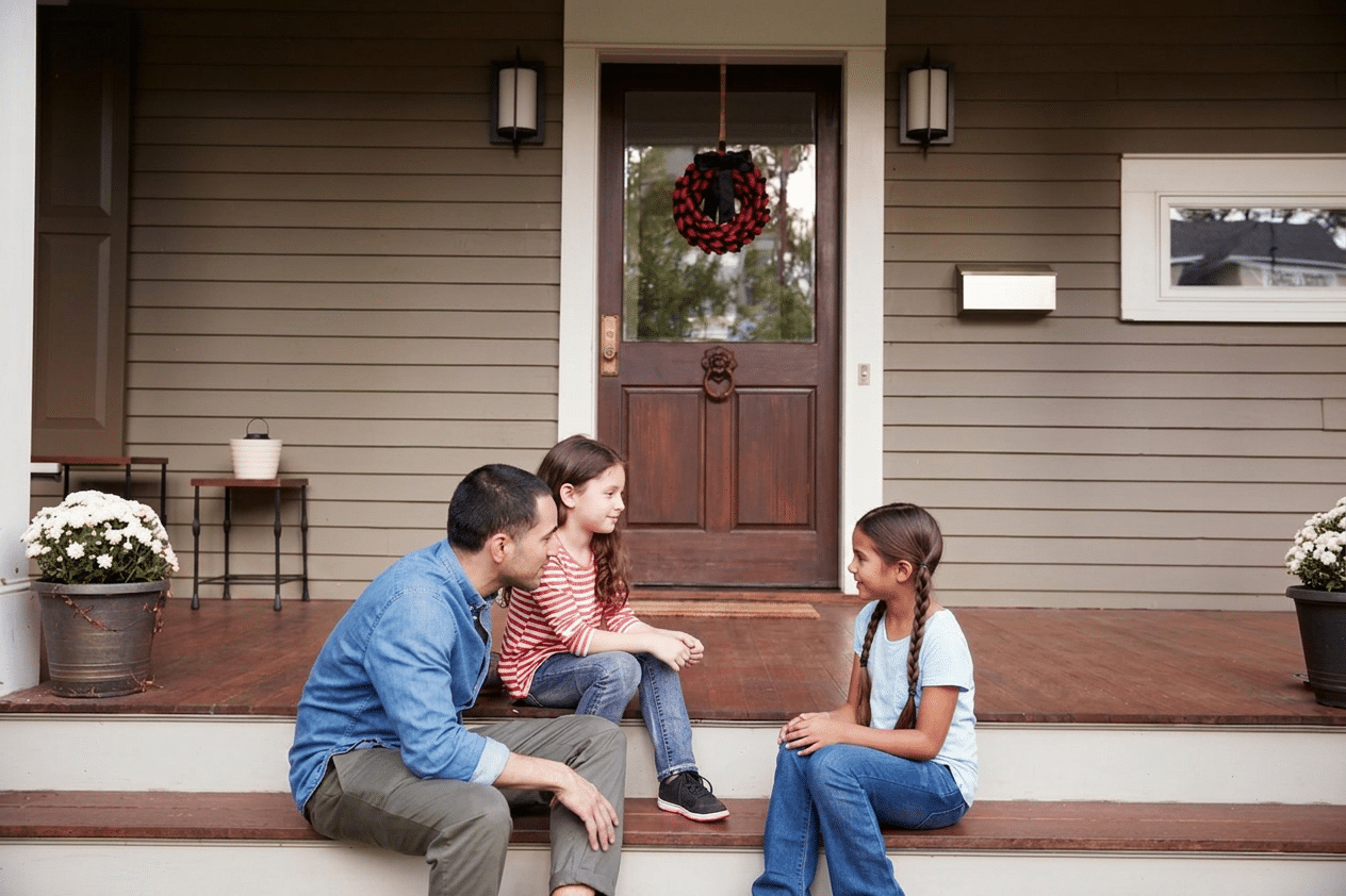 Father and two daughters sitting on front porch steps in front of a wood entry door — residential entry door replacement in Utah and Boise