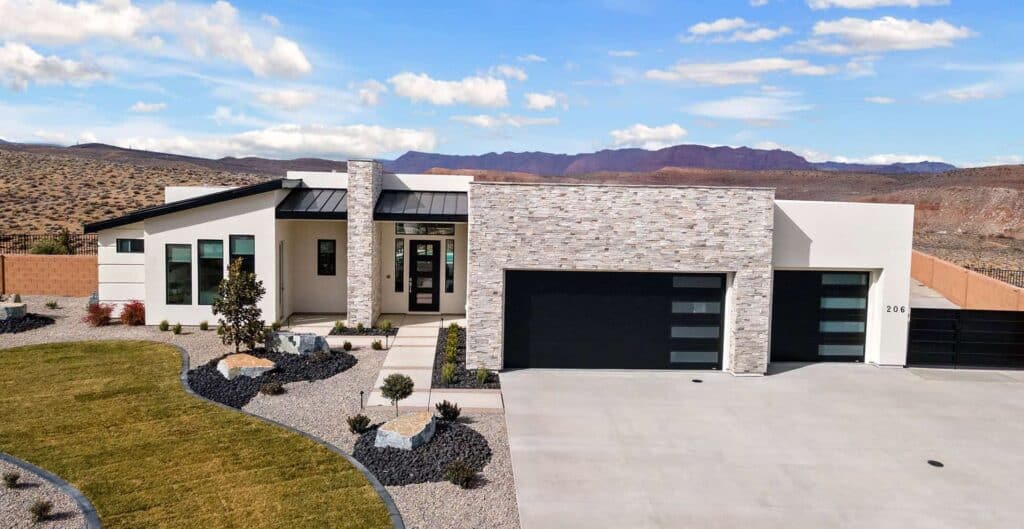 Modern home with two matte black steel and glass garage doors, white stucco and ledger stone exterior, and red rock desert mountains in the background, typical of contemporary construction in St. George, Utah