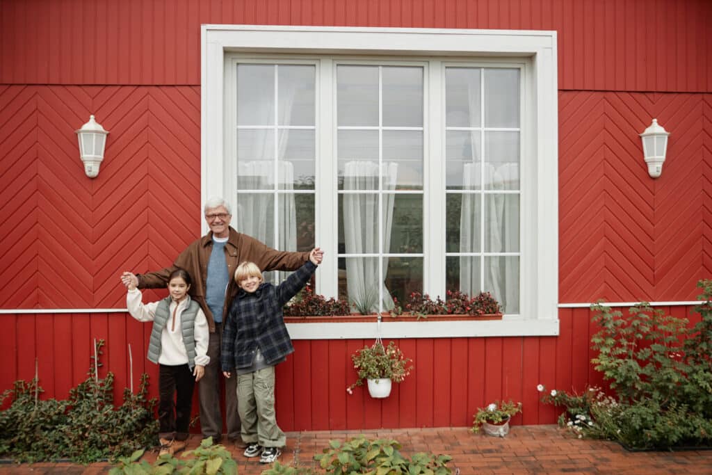 Grandfather and two grandchildren standing in front of large white triple-pane windows on a red home