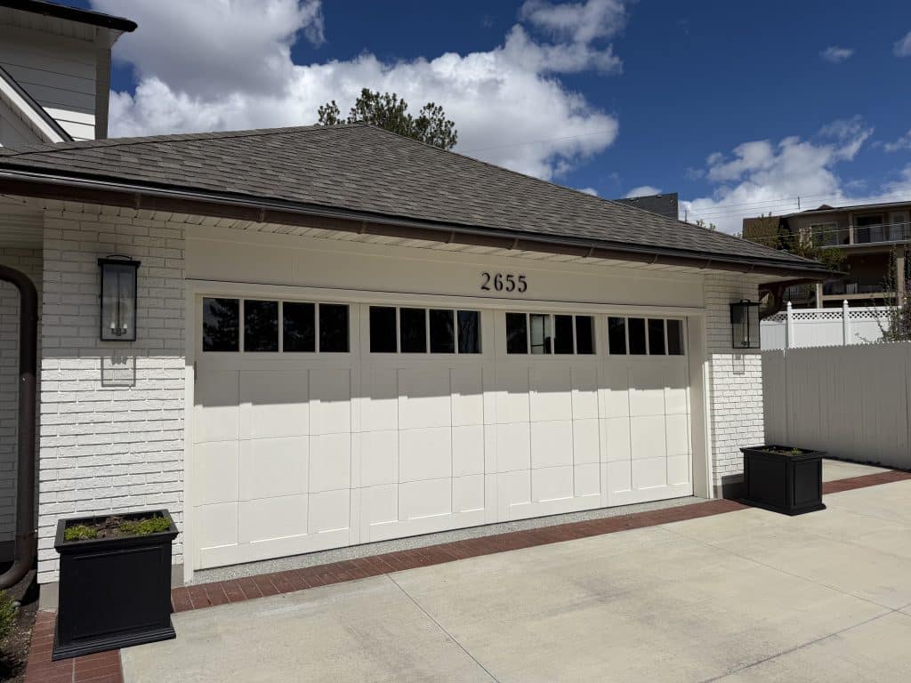 Cream-colored carriage-style steel garage door with a row of rectangular windows across the top, marked with the house number 2655, installed on a white painted-brick home with a brown shingled roof and black coach lights.