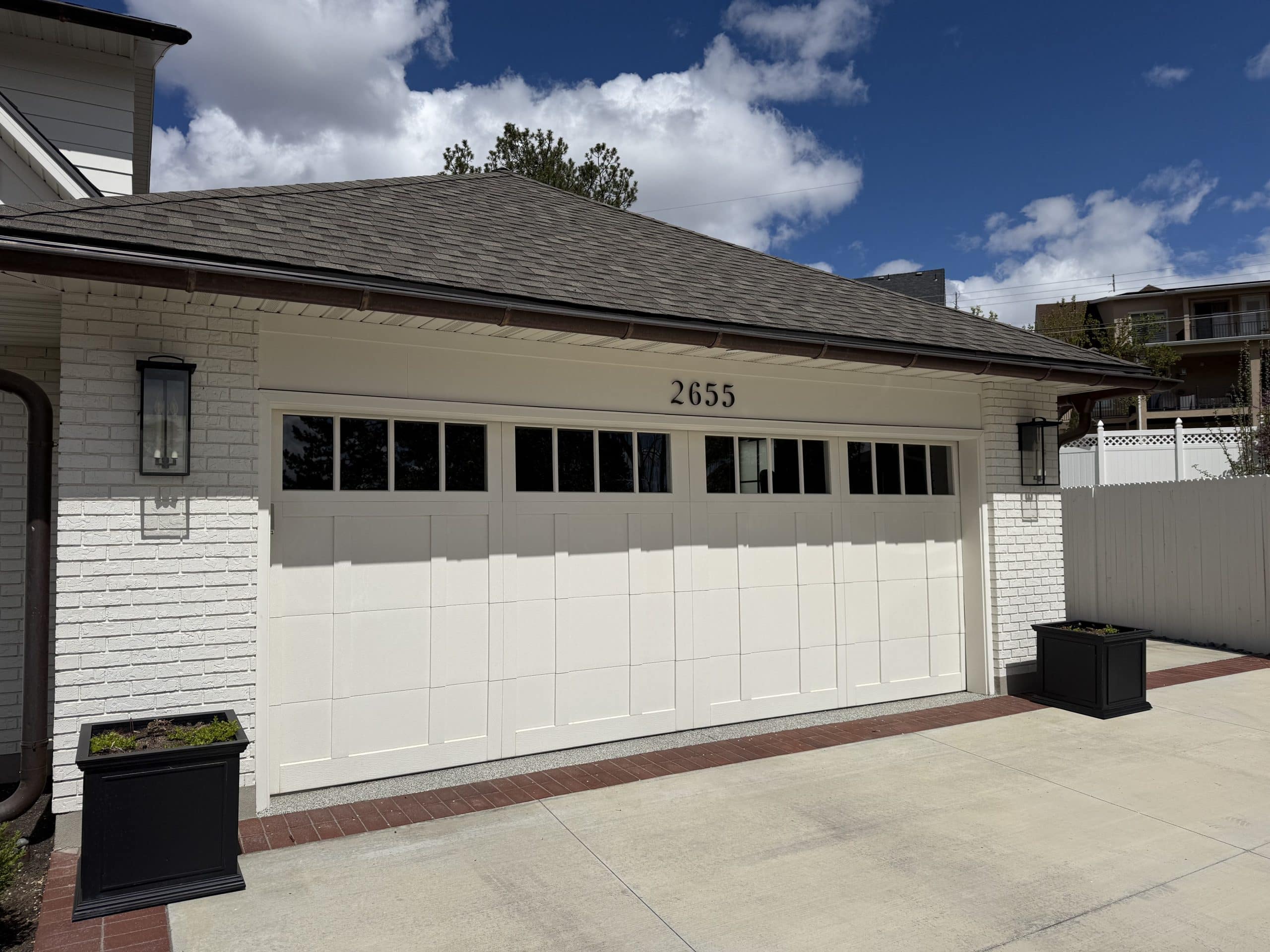 Cream-colored carriage-style steel garage door with a row of rectangular windows across the top, marked with the house number 2655, installed on a white painted-brick home with a brown shingled roof and black coach lights.